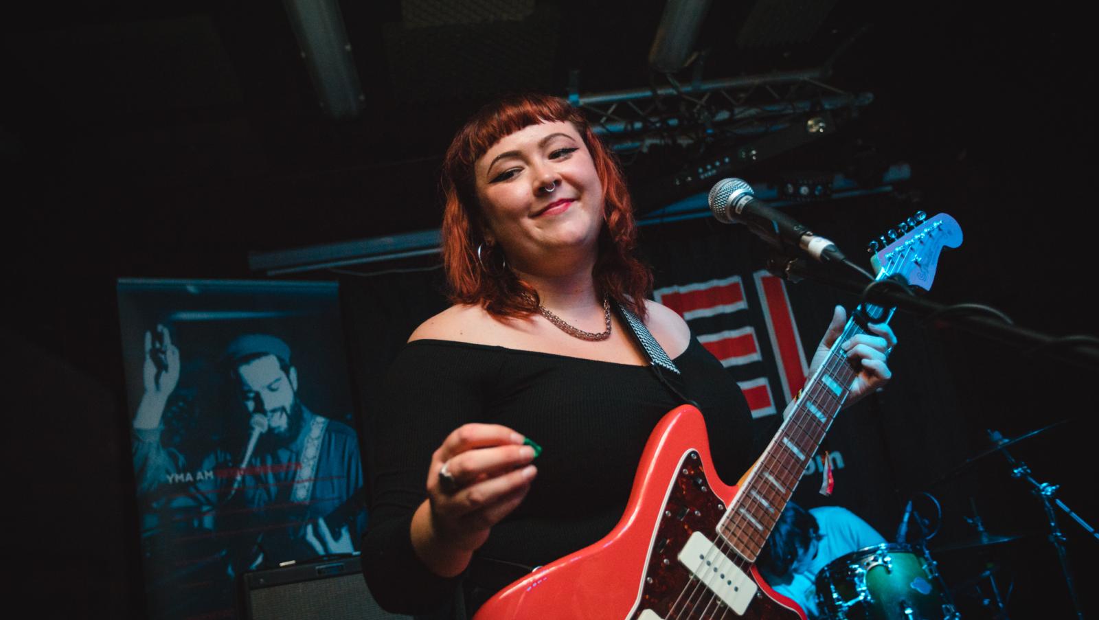 Close of up lead singer from Adwaith, Hollie Singer. Hollie has a short red bob and is wearing a black off the shoulder top. She is also holding a red electric guitar.