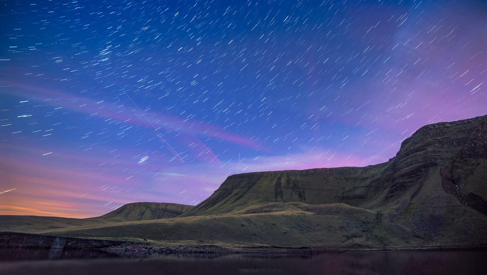 Outline of lake and mountain backdrop against a dark blue and purple night sky, dotted with blurry stars