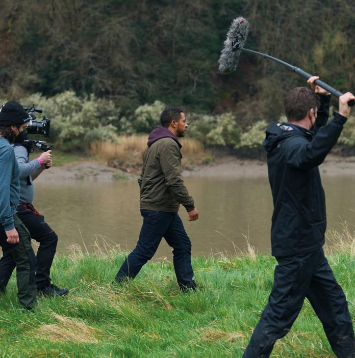Four people filming next to a river. One person is in shot, while the other three follow – one holds a boom and the other a camera.