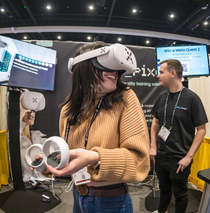 A woman wearing a peach-coloured jumper stands in front of two screens while wearing a virtual reality headset.