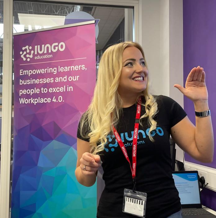 A blonde-haired woman stands, smiling in front of a purple, blue and pink iungo banner. 
