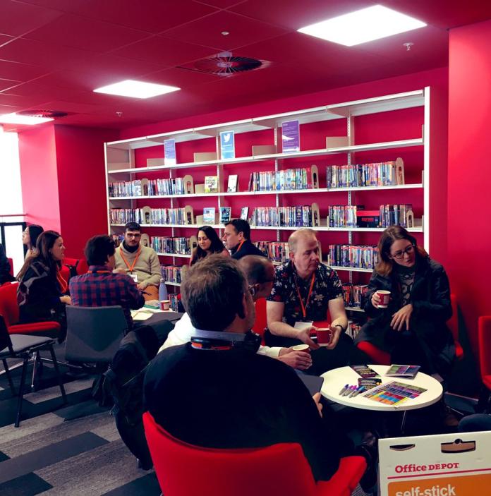 Medium shot of group of people sitting and talking in a red room with a wall lined with books.