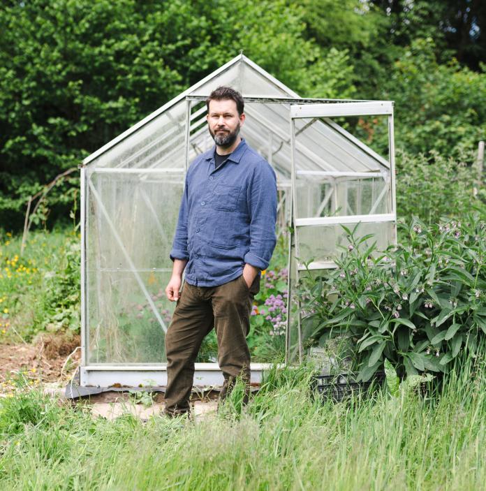 Full length shot of a person wearing a blue shirt and brown trousers standing in front of a greenhouse in a garden.