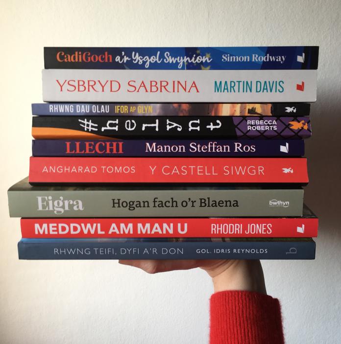 A hand wearing a red knit holds a stack of nine different coloured books against a white wall.