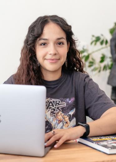 Close up shot of Yassmine using her Macbook. Yassmine has long brown curly hair and is wearing a grey tshirt with a animated graphic.