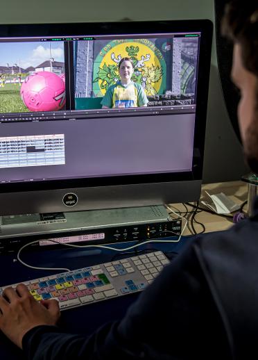 Over the shoulder shot of Seb Jones editing videos on a monitor. The screen shows a split screen image of two stills – one with a close up of a person against a green and yellow background. The other with a large pink ball in a grassy field. 