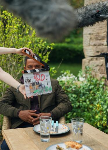 Two people on set in front of the camera against a backdrop of greenery. One is seated at a wooden table. The other is holder a movie clap board.