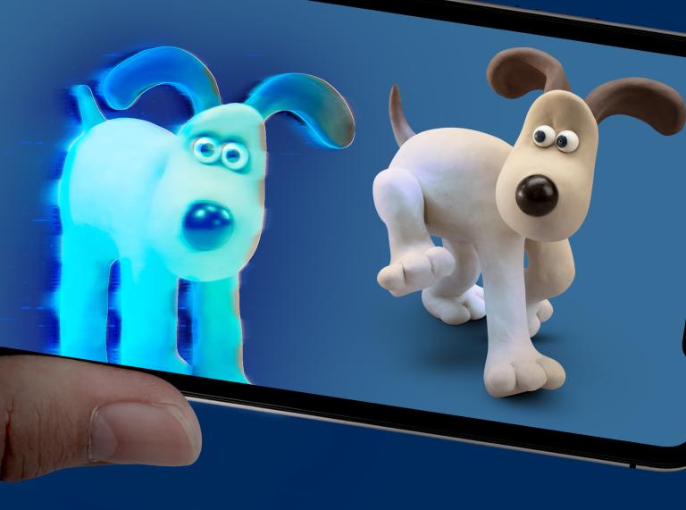 Close up of hand showing a phone screen with Gromit from Wallace and Gromit against a blue background. Gromit is a white animated dog with a black nose and brown ears. He's standing with one leg up next to a blue AR version of himself.