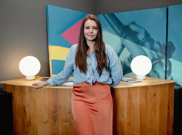 Medium shot of Robyn standing in front of a wooden desk at Bomper Studios in Cardiff. Robyn is wearing an orange midi shirt and a blue shirt. The wall behind her is covered in blue, yellow and pink graphic patterns.