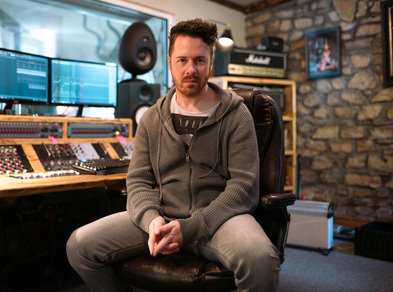 Mid-shot of Jayce Lewis of Northstone Studios sitting facing the camera in his studio. Jayce wears grey jeans and a grey zipped hoodie.