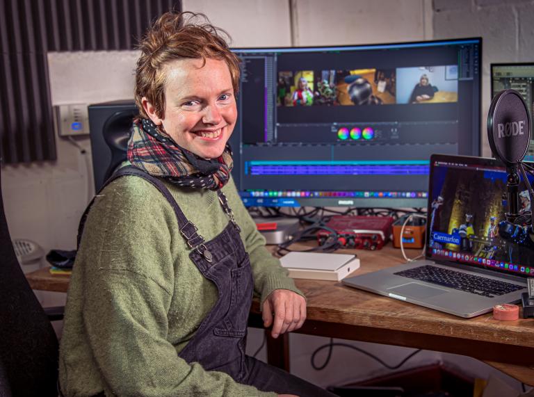 Person wearing dungarees over a green jumper with a check scarf sits at a desk with a laptop, monitor screen and microphone.
