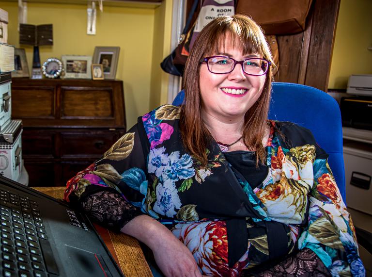 Medium shot of Rebecca Roberts. Rebecca is sitting at her desk at home in a yellow room. She's wearing a black dress with a colourful flower print, rectangle dark red glasses and has medium-length brown hair.
