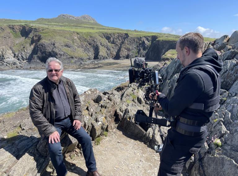 One person stands holding a film camera pointed at another person sitting on an old stone wall. The backdrop show the sea against a grass-topped cliff.