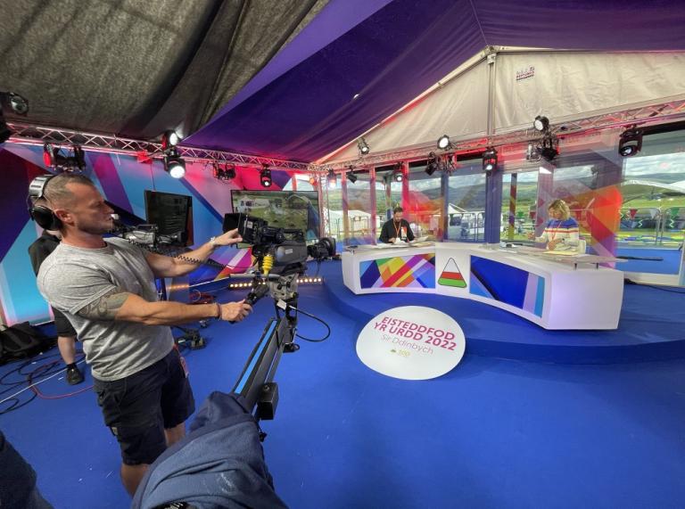 A person operates a film camera in a studio with blue carpets, a white desk and a sticket on the floor saying Eisteddfod Yr Urdd 2022.