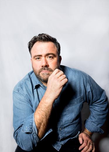 Close up portrait shot of Celyn Jones. Celyn is wearing a blue navy shirt and is sitting against a plain grey background. They have short brown hair and short brown facial hair.