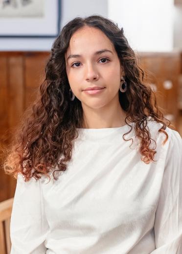 Close up shot of Yassmine. Yassmine is wearing a long white top and has curely brown hair.