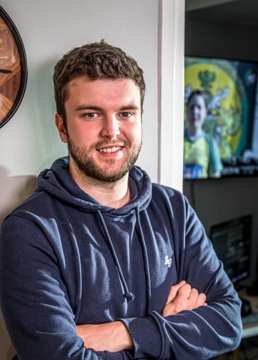 Close up shot of Seb Jones. Seb is sitting with their arms crossed wearing a blue hoodie. They have dark blonde hair and short facial hair around the mouth and nose.