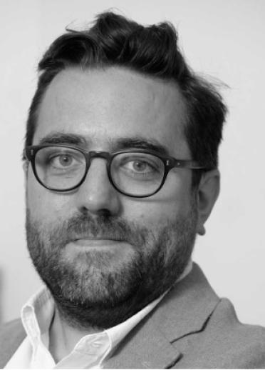 Close up, grey-scale shot of Dan Cheesbrough. Dan has short brown hair, a beard, and wears thin brown glasses.