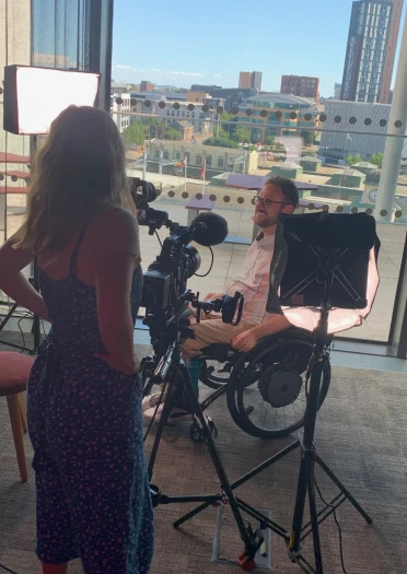 Man in a wheelchair being filmed in a bright room with cityscape views in the background