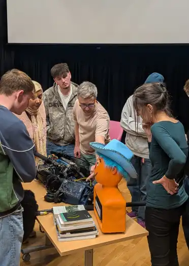 Group of people gathered around equipment and an orange mannequin with a blue hat on a table