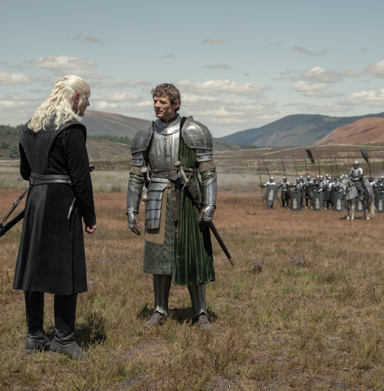 Two armored figures facing each other on a grassy field with soldiers and hills in the background