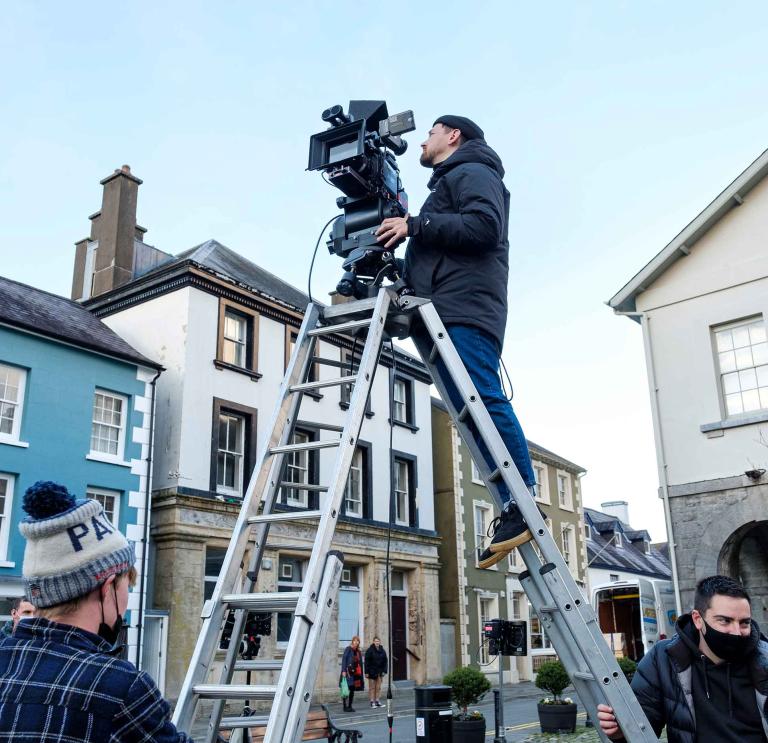 Surrounded by three-story buildings, a person stands at the top of tall step ladder to film with a camera