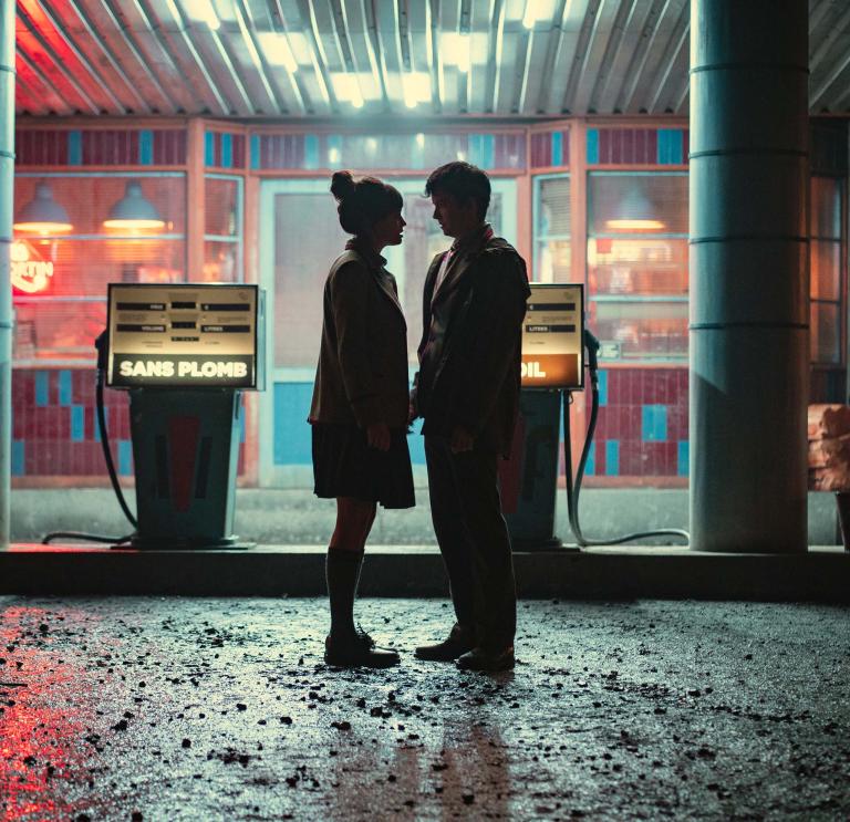 Shadow outline of Emma Mackey as Maeve Wiley and Asa Butterfield as Otis Milburn standing close together in front of a retro petrol station at night