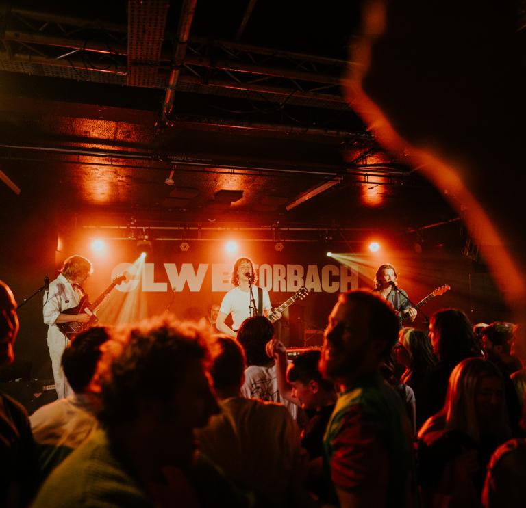 A busy crowd under red lights at a Buzzard Buzzard Buzzard gig at Clwb Ifor Bach