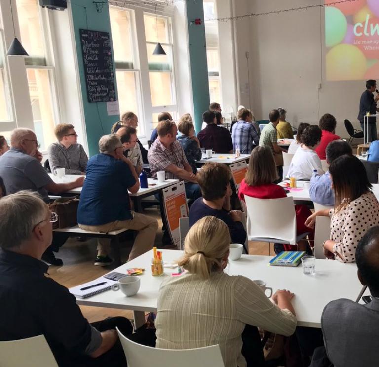 A large group of people sitting at white tables facing a projector screen as someone presents. The screen shows a background of colourful balloons with the word "Clwstwr" in white text in the middle.