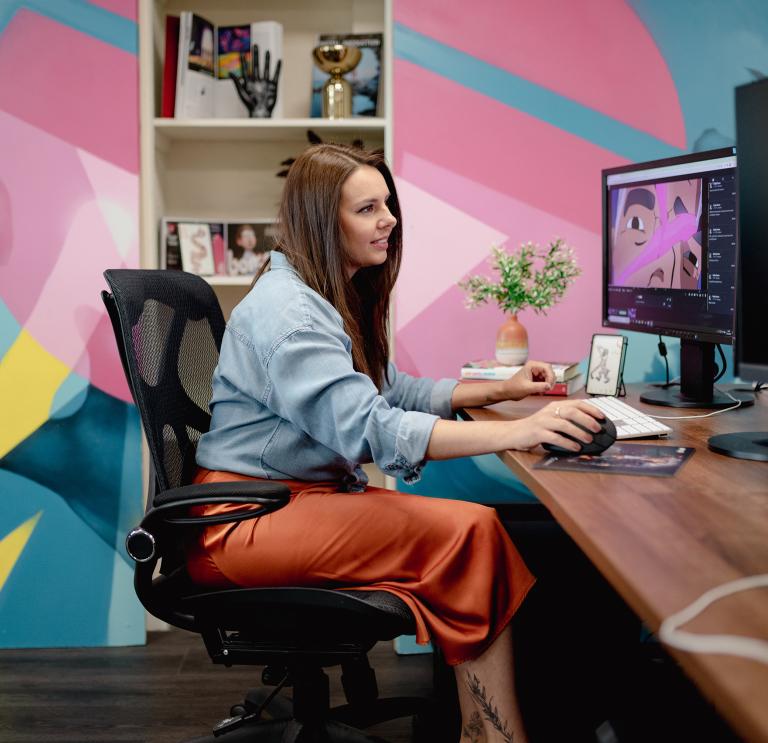 Medium shot of Robyn working at her wooden desk at Bomper Studios in Cardiff. Robyn is wearing an orange midi shirt and a blue shirt. The wall behind her is covered in blue, yellow and pink graphic patterns. 