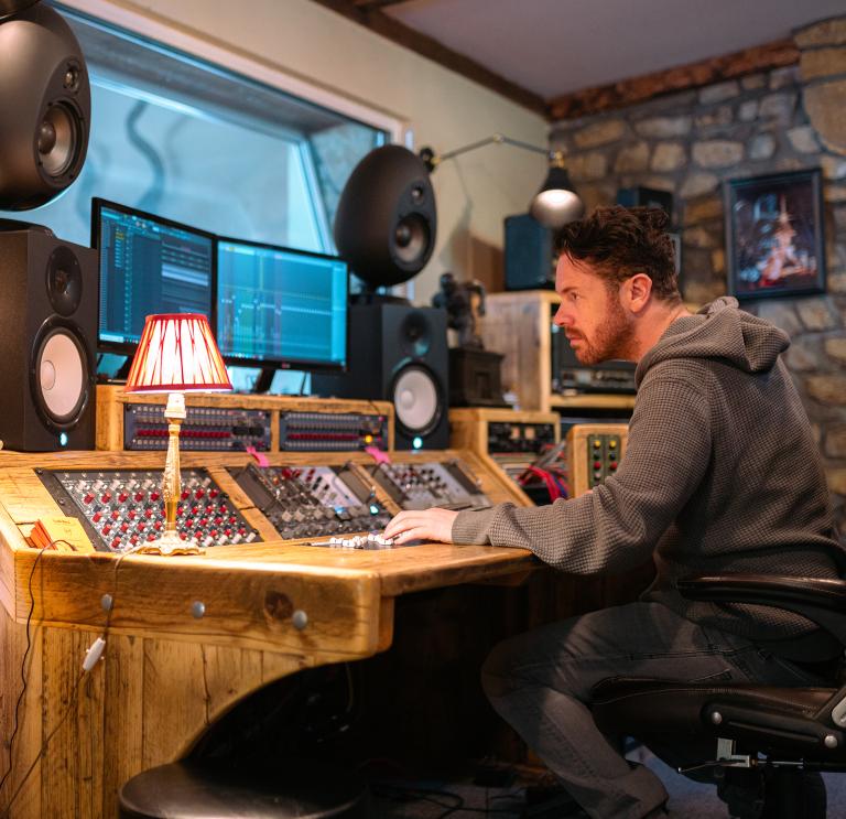 Jayce Lewis of Northstone Studios sits at a wooden mixing desk with monitors and large speakers in a dimly-lit studio. 