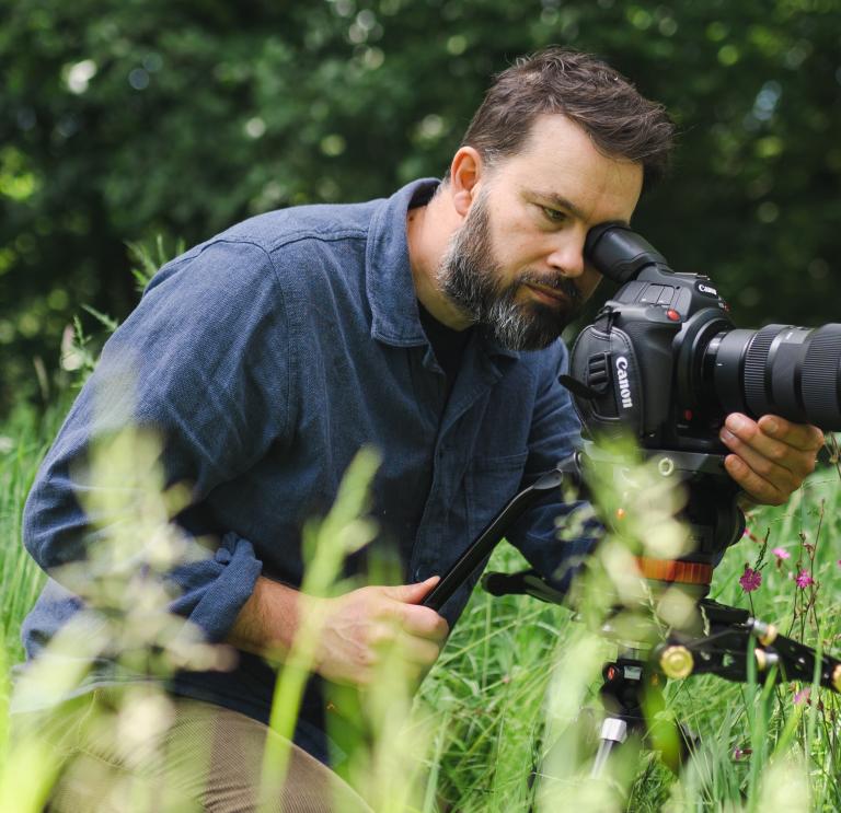 Close up shot of a person wearing a blue coloured shirt looking through a camera lens.