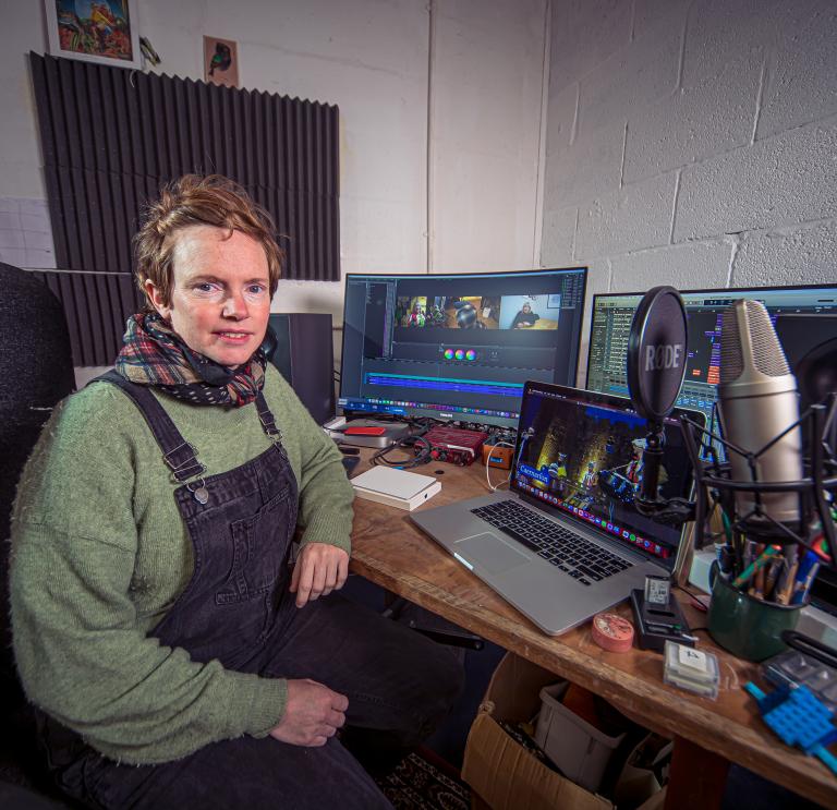Person wearing dungarees over a green jumper with a check scarf sits at a desk with a laptop, monitor screen and microphone.