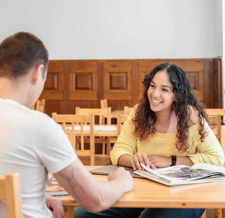 Close up shot of Yassmine from Bomper Studio talking to colleague. Yassmine is smiling, sitting at a wooden table wearing a yellow of the shoulder top.