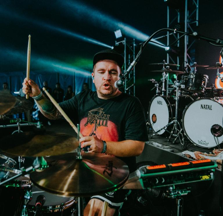 Medium shot of Ryan Richards playing drumps on a dimly lit stage. Ryan is wearing a black baseball cap and a black tshirt with an orange design. 