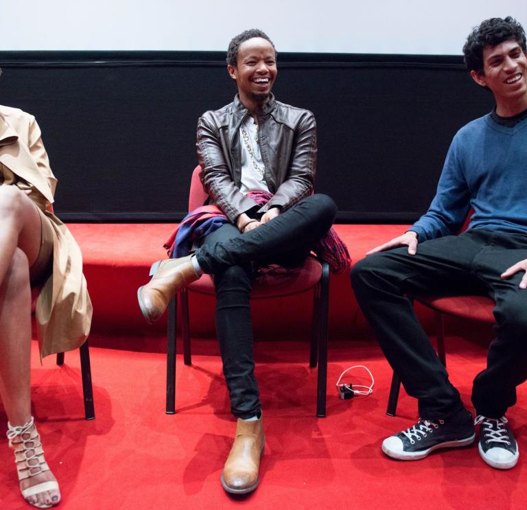 Three people sit together on red chairs. They are all looking to towards the right of the shot and laughing. The floor is red carpet and the wall behind is half black and half white. 