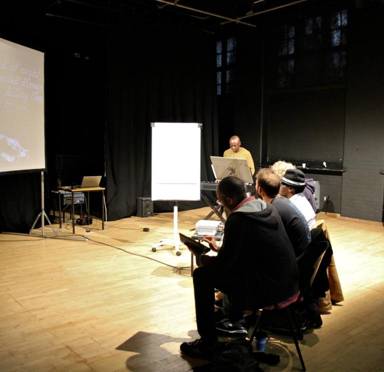 Group of people sit in a dimly lit hall surrounding by black walls. There is a person standing at the front talk. Next to them is a white paper flipboard. 