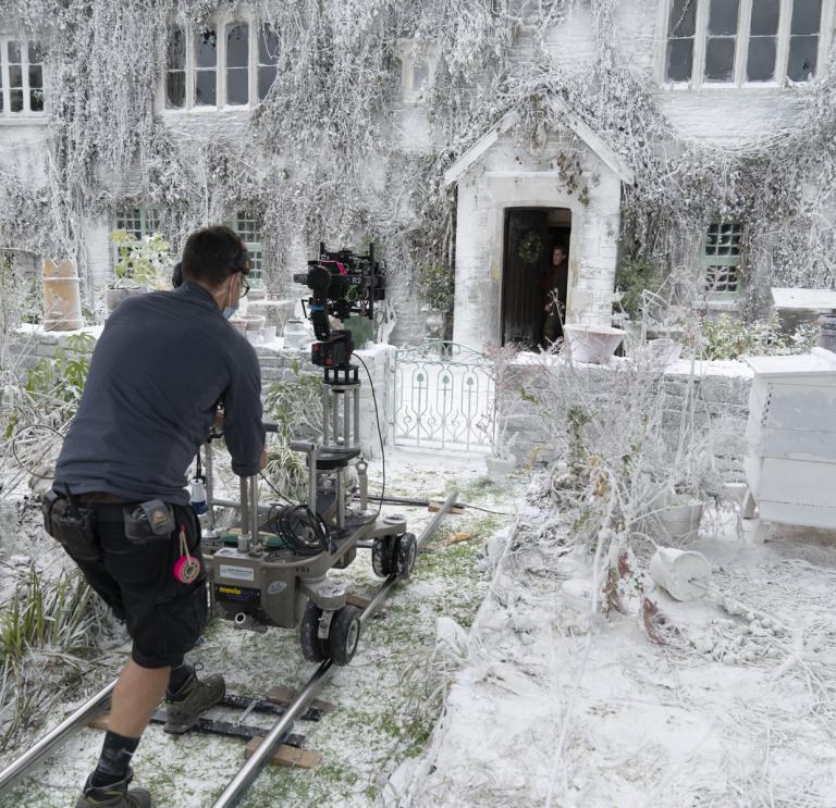 A person stands on a mini track holding a film camera. They are focusing on a white house covered in snow.