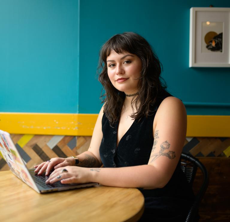Yasmine has dark brown hair and sits at a table on a laptop and smiles at the camera