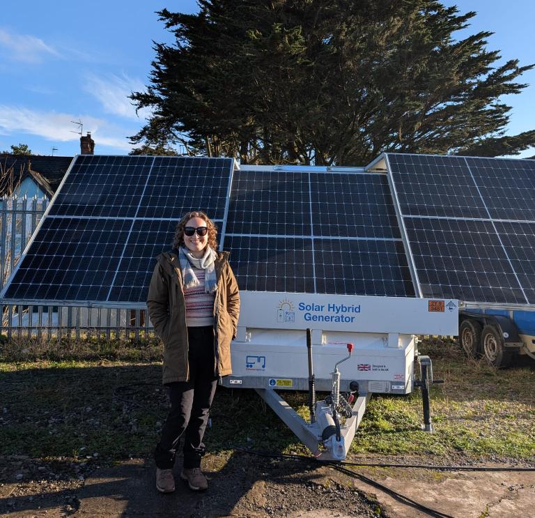 Person standing in front of a solar hybrid generator with solar panels and a tree in the background