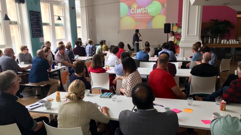 A large group of people sitting at white tables facing a projector screen as someone presents. The screen shows a background of colourful balloons with the word "Clwstwr" in white text in the middle.