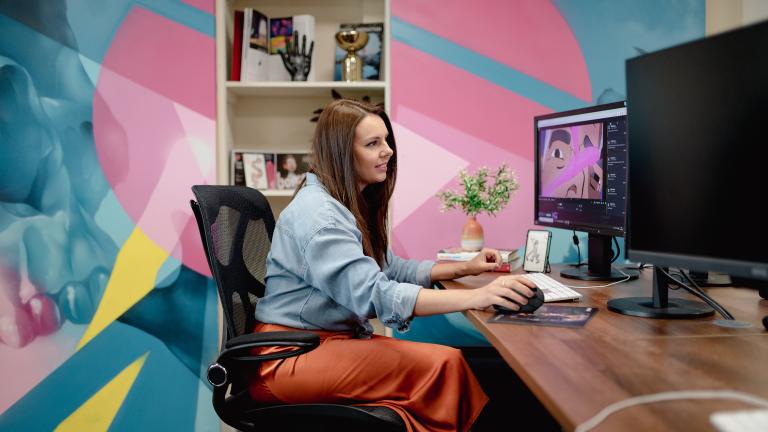 Medium shot of Robyn working at her wooden desk at Bomper Studios in Cardiff. Robyn is wearing an orange midi shirt and a blue shirt. The wall behind her is covered in blue, yellow and pink graphic patterns.