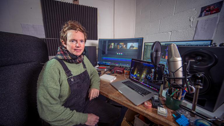 Person wearing dungarees over a green jumper with a check scarf sits at a desk with a laptop, monitor screen and microphone.