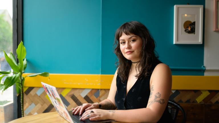 Yasmine has dark brown hair and sits at a table on a laptop and smiles at the camera
