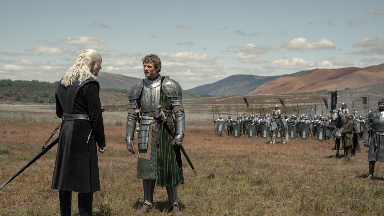 Two armored figures facing each other on a grassy field with soldiers and hills in the background