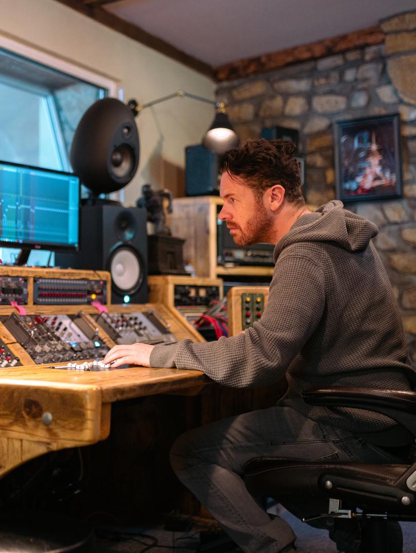 Jayce Lewis of Northstone Studios sits at a wooden mixing desk with monitors and large speakers in a dimly-lit studio. 