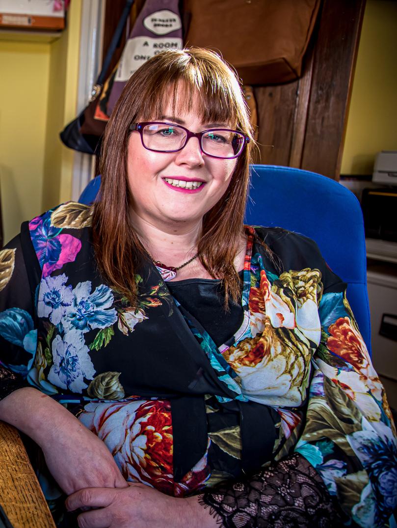 Medium shot of Rebecca Roberts. Rebecca is sitting at her desk at home in a yellow room. She's wearing a black dress with a colourful flower print, rectangle dark red glasses and has medium-length brown hair.