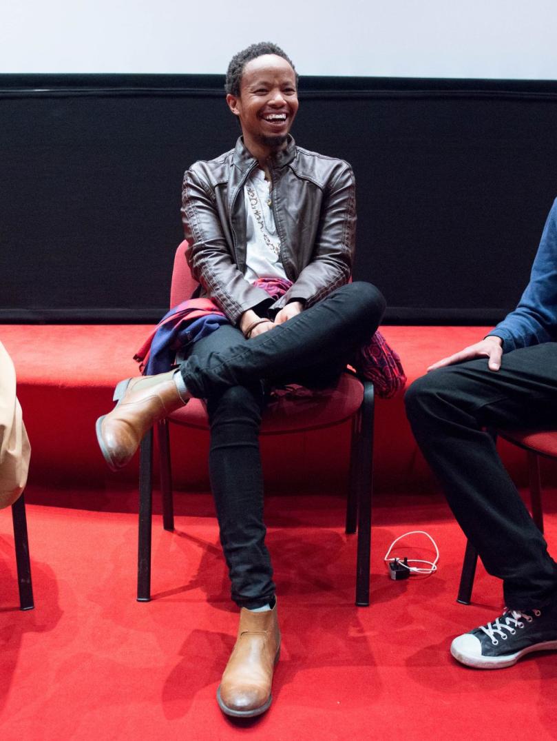 Three people sit together on red chairs. They are all looking to towards the right of the shot and laughing. The floor is red carpet and the wall behind is half black and half white. 