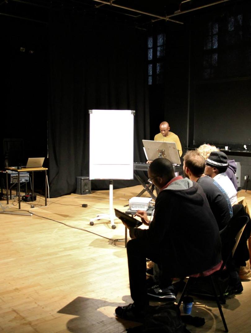 Group of people sit in a dimly lit hall surrounding by black walls. There is a person standing at the front talk. Next to them is a white paper flipboard. 