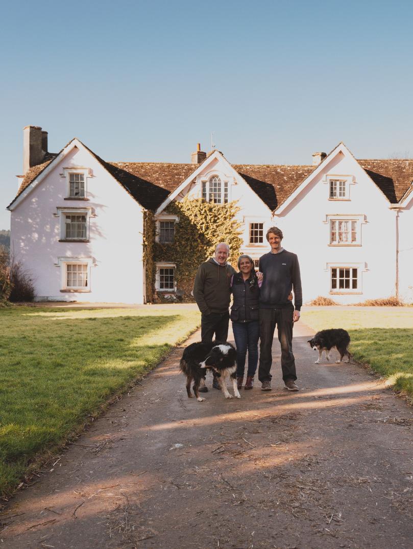 A man standing with a woman and a younger man in front of a large pink farm house on a sunny day